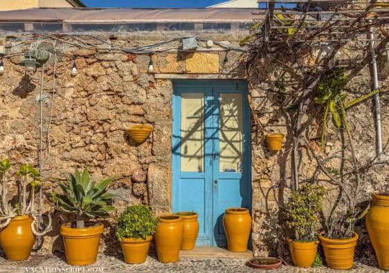 turquoise front door with orange potted plants lining the ground of this Sicilian front door