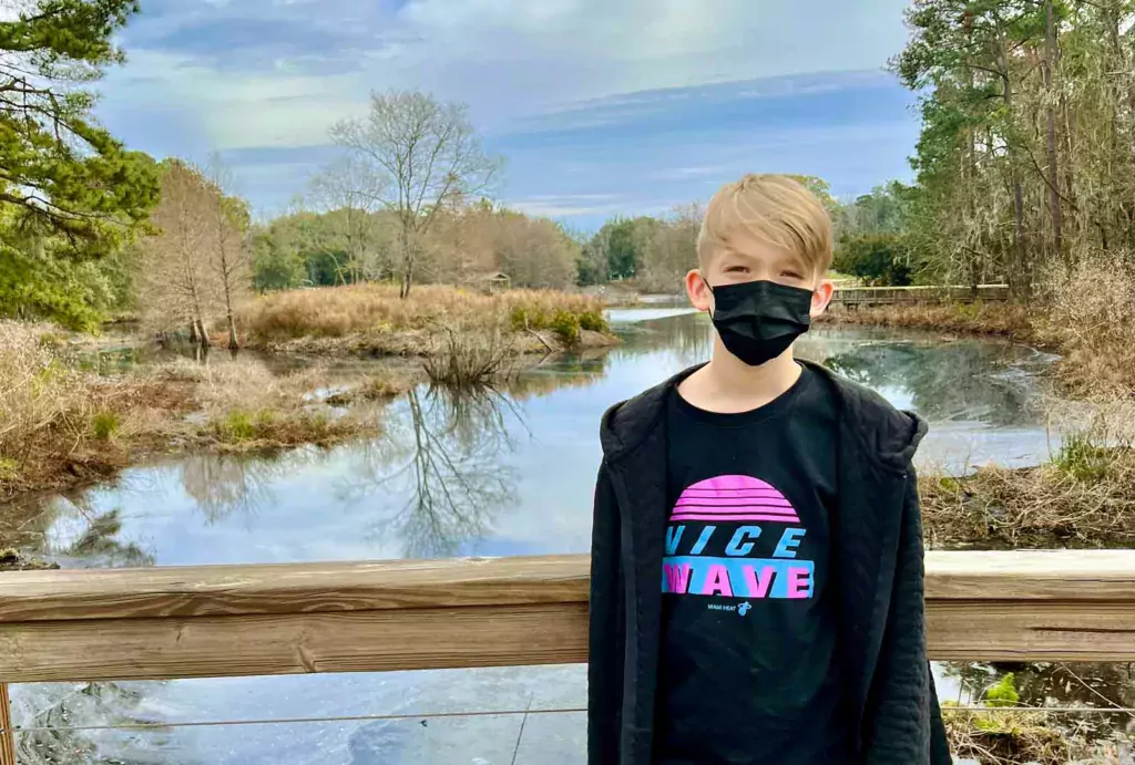 A boy wearing a mask in front of the Cypress Wetlands boardwalk, overlooking the water during winter with brown-tinged bushes.