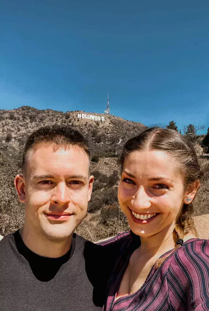A couple delighting in their visit to the Hollywood Sign, captured from a lower vantage point with the iconic sign towering in the background.