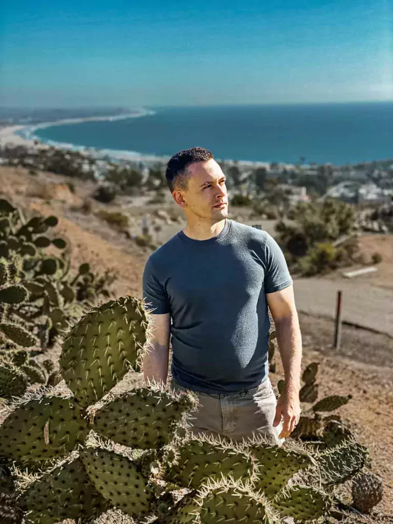 A striking collection of cacti surrounds a buff man as he gazes toward the sun, with the curving coastline in the background.