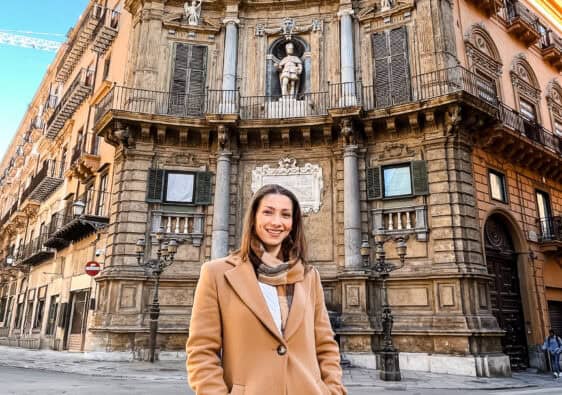 Smiling woman in a brown trench coat standing in front of Quattro Canti, a historic Baroque corner in Palermo, Sicily.