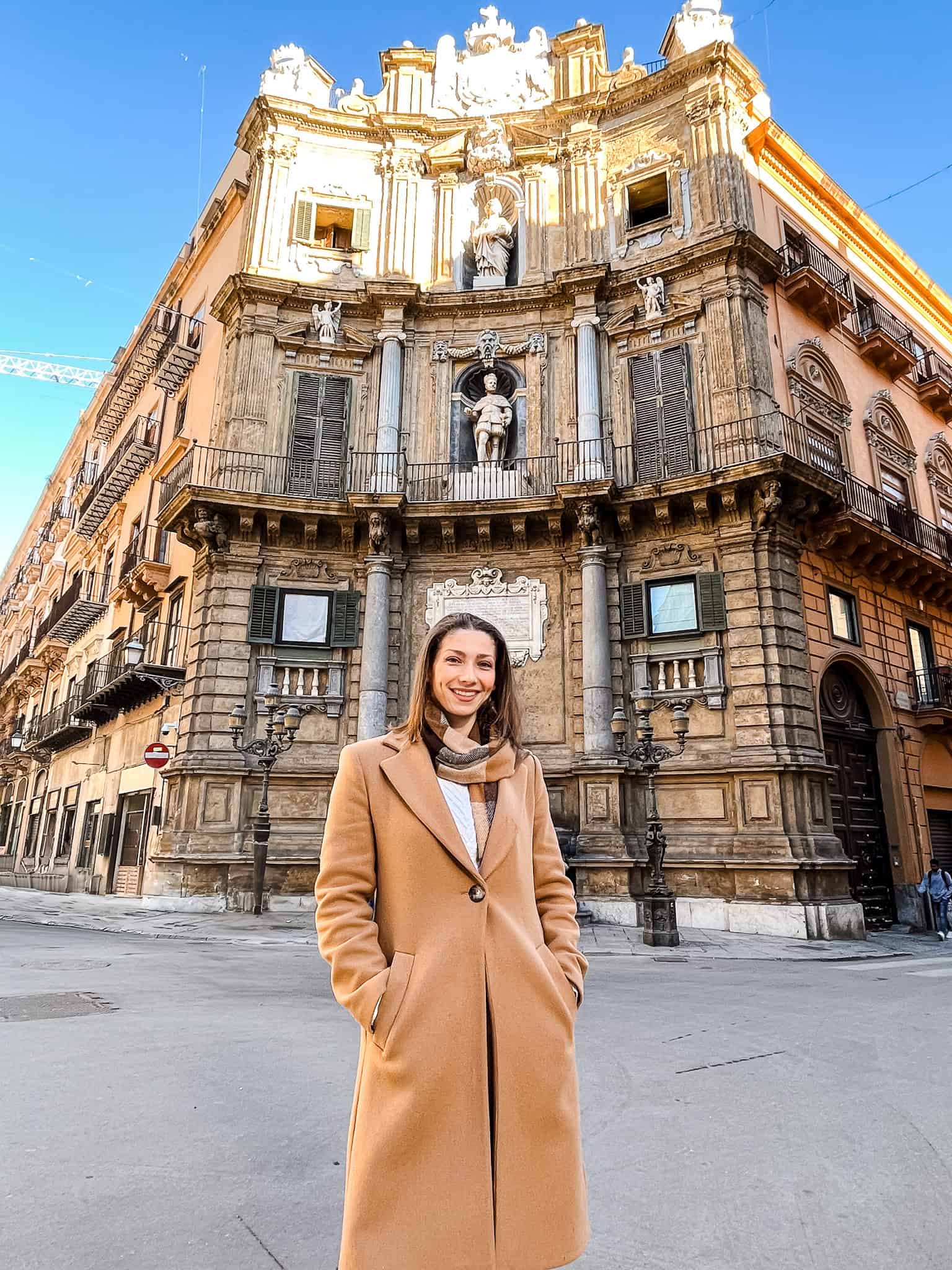 Smiling woman in a brown trench coat standing in front of Quattro Canti, a historic Baroque corner in Palermo, Sicily.