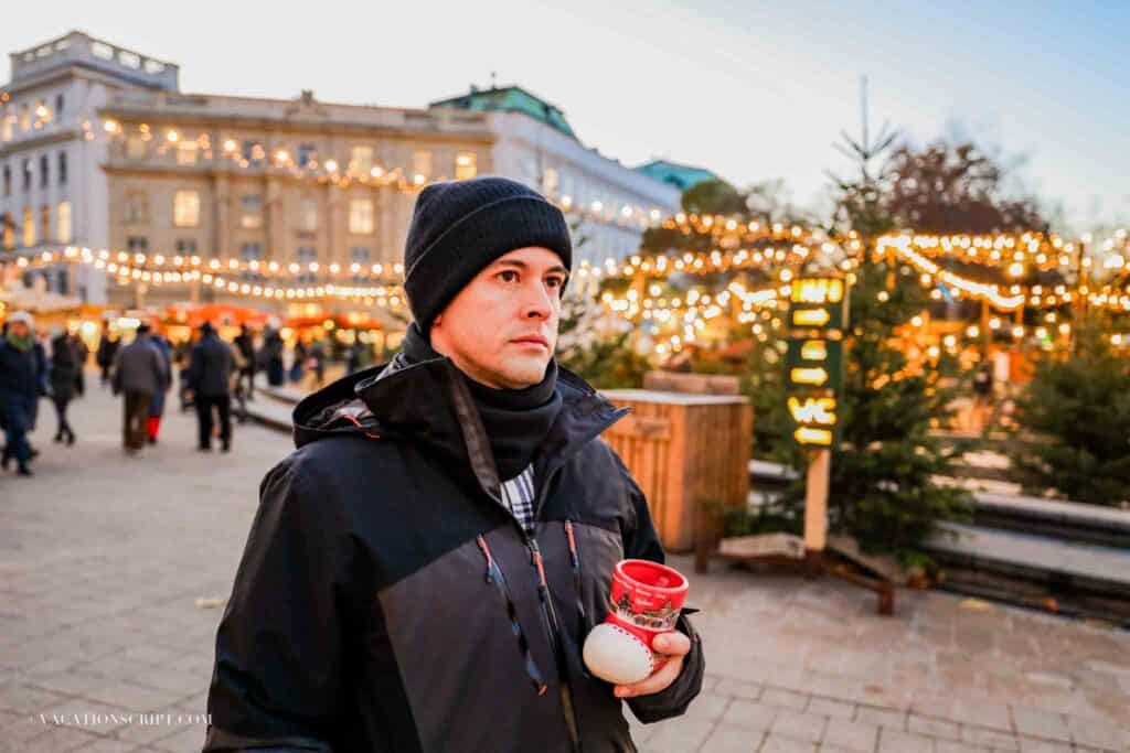 handsom man holding apple rum drink at Karlsplatz market with lights and christmas trees behind him