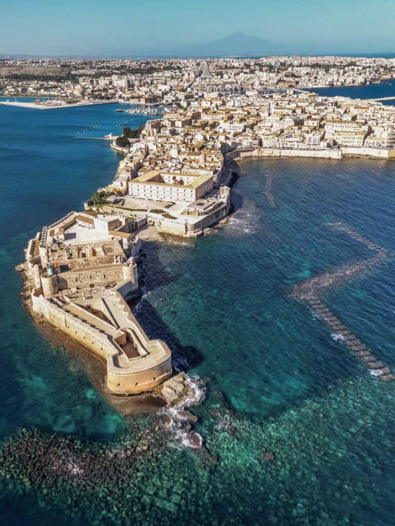 Aerial view of Castello Maniace in Ortigia, Sicily, with the fortress on a peninsula surrounded by turquoise blue sea and visible submerged stone steps.