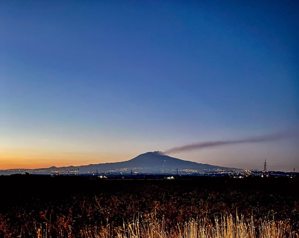 Mount Etna at twilight with a smoke line drifting to the east showing the city lights up the volcano.