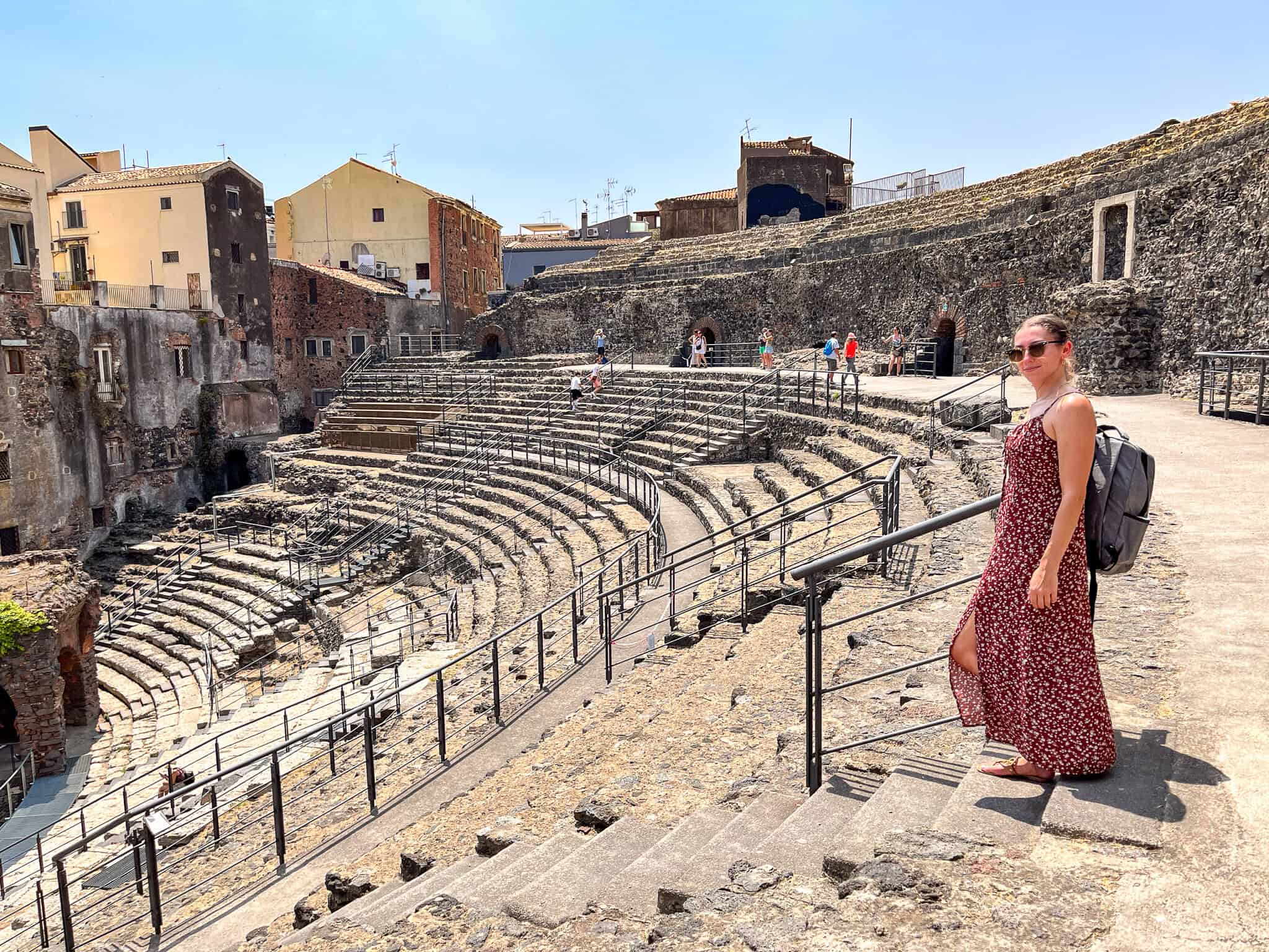 Woman in a red floral dress with a backpack and sunglasses walking down the ancient stone steps of the Roman-Greek Theater in Catania on a bright, sunny day.
