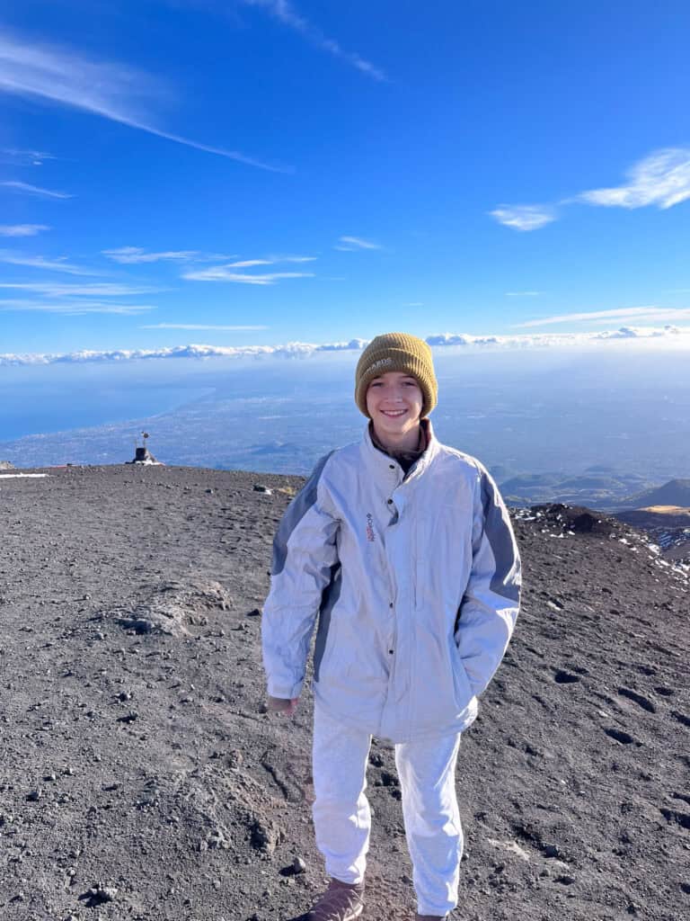 Smiling boy standing on black ash-covered ground at the summit of Mt. Etna, with clouds, blue sky, and a distant view of Catania below.