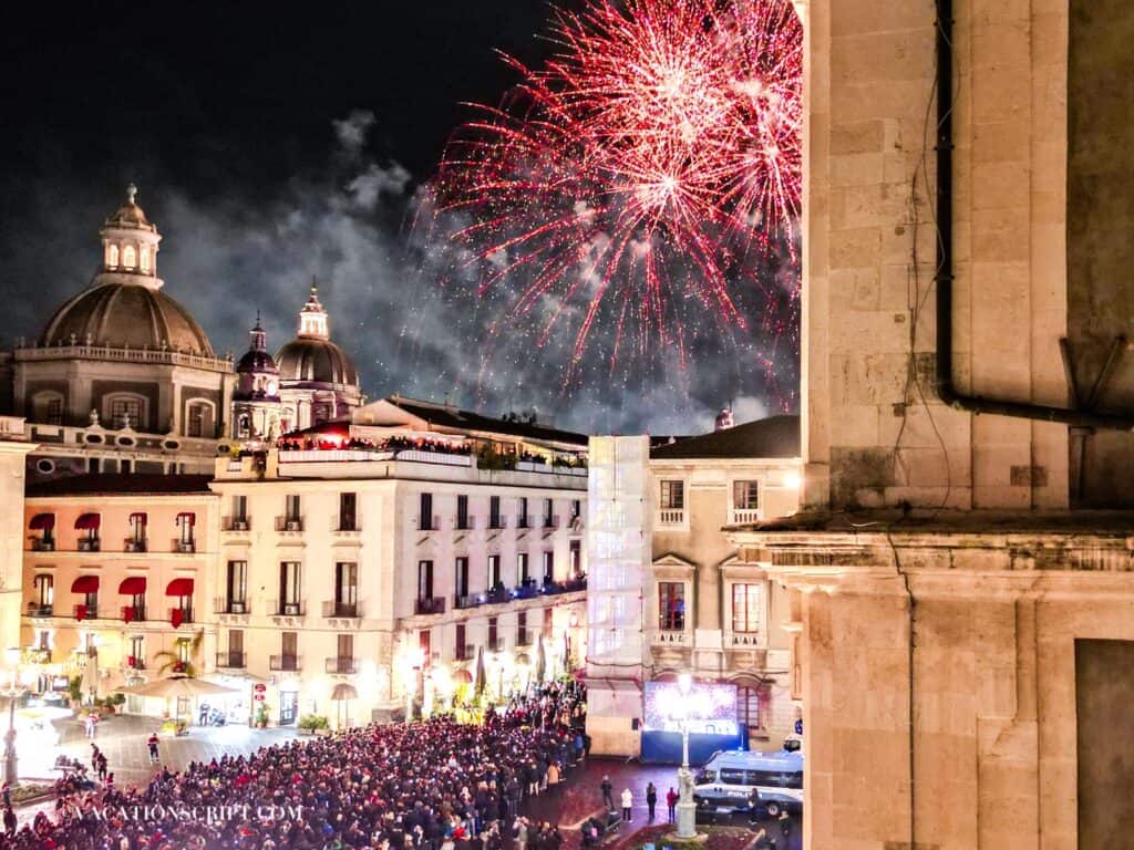 Nighttime view from a hotel balcony overlooking Piazza Università in downtown Catania, with red fireworks lighting up the sky and a large crowd gathered to watch for the annual Sant’Agata Festival.