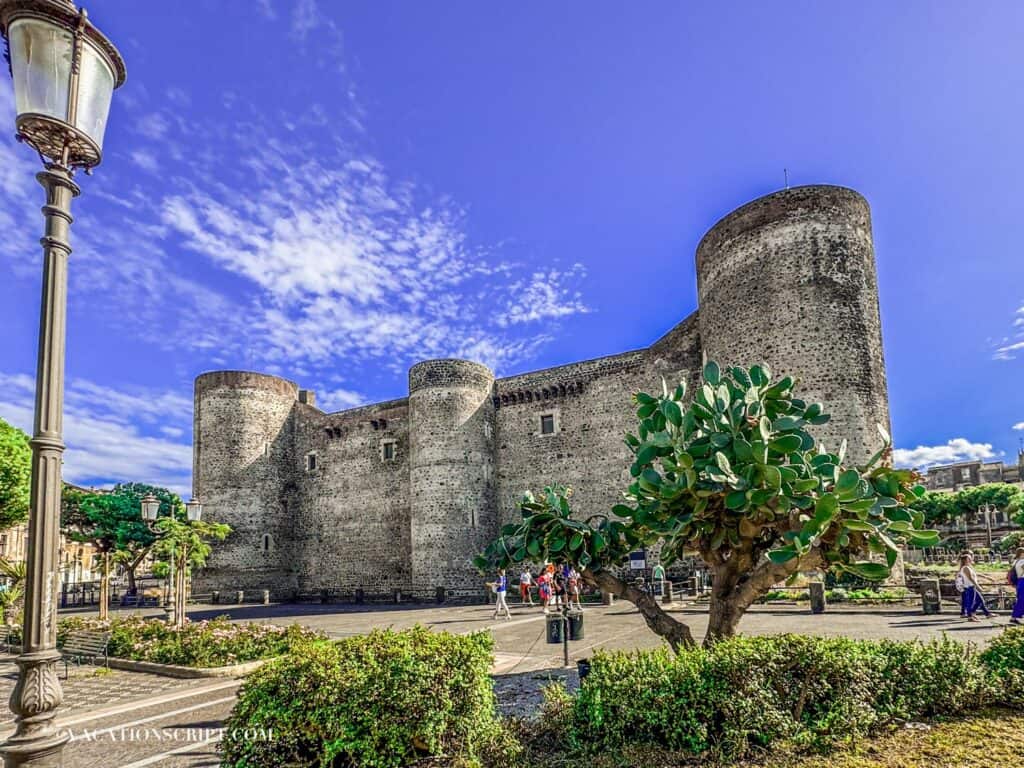 Ursino Castle in Catania, Sicily on a sunny day, with a towering cactus tree, cobblestone path, vintage street lamp, and locals strolling nearby.