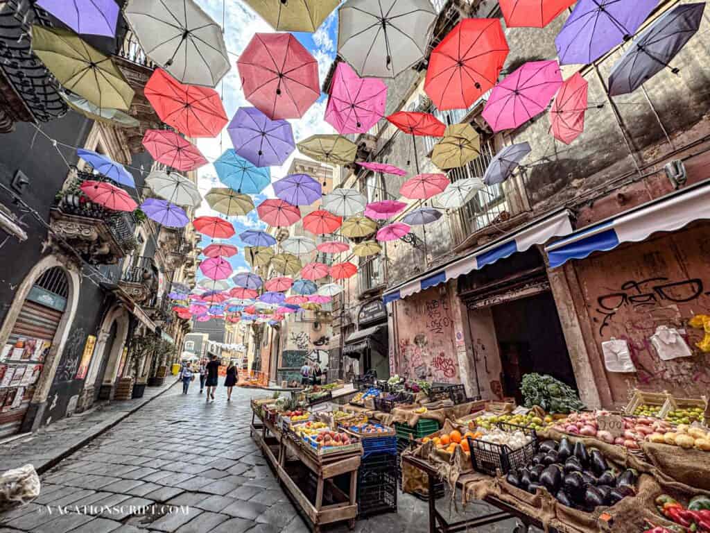 Colorful umbrellas suspended above a lively alley in Catania’s fish market, Sicily, with a fruit and vegetable vendor’s stall ready for the day.