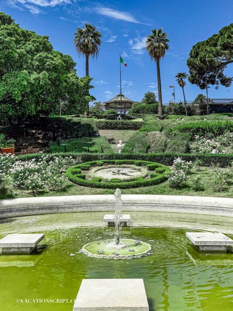 View from the fountain at Bellini Gardens in Catania, showing a hillside pavilion with a clock shaped from bushes, a statue, colorful flowers, and the daily updated date displayed in front of an Italian flag.