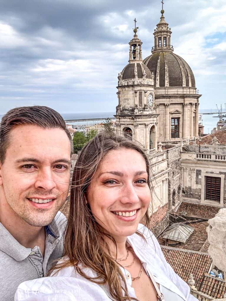 Smiling couple at the top of the Church of the Abbey of Sant'Agata in Catania, with panoramic views of the city and the Cathedral of Sant'Agata in the background.