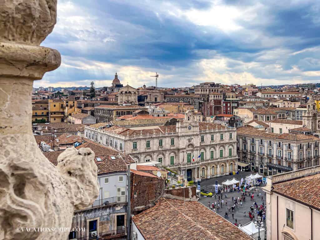 Bird’s-eye view of Piazza Università in Catania from the top of the Church of the Abbey of Sant’Agata, with dramatic sun rays breaking through clouds and illuminating locals walking below.